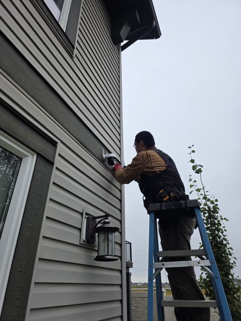 Technician on a ladder installing an outdoor security camera on the siding of a Calgary house